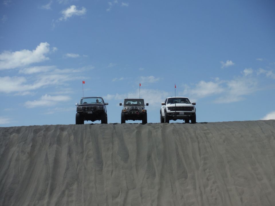 Three off-road vehicles, including a white Ford F-150 Raptor and a dark Jeep Wrangler, are lined up on a sand dune under a blue sky with clouds