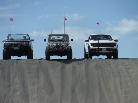 2005 Jeep Wrangler - Three off-road vehicles, including a 2005 Jeep Wrangler, are lined up on a sand dune under a blue sky.