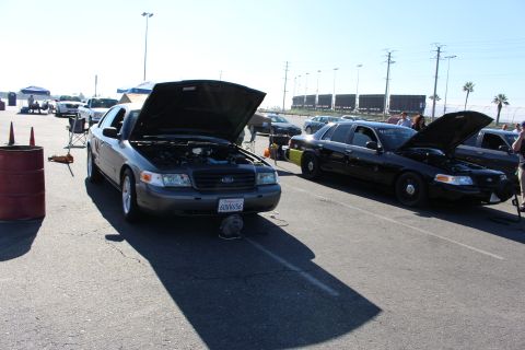 2003 Ford Crown Victoria - Two dark Ford sedans sit with hoods raised in a paved lot, the nearer car showing a California plate and Ford badge.