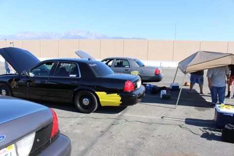 2003 Ford Crown Victoria - Black sedans with raised hoods and windshield number decals sit at a track staging lot, a canopy and coolers nearby, mountains beyond.