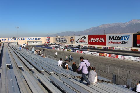 2003 Ford Crown Victoria - People sit on aluminum grandstands overlooking the Auto Club Dragway strip, with Lucas Oil and MAVTV banners and mountains beyond.
