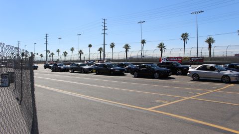 2003 Ford Crown Victoria - Dark coupes and sedans stage in a line behind a chain-link fence in a paved track lot with palm trees and a Lucas Oil banner.