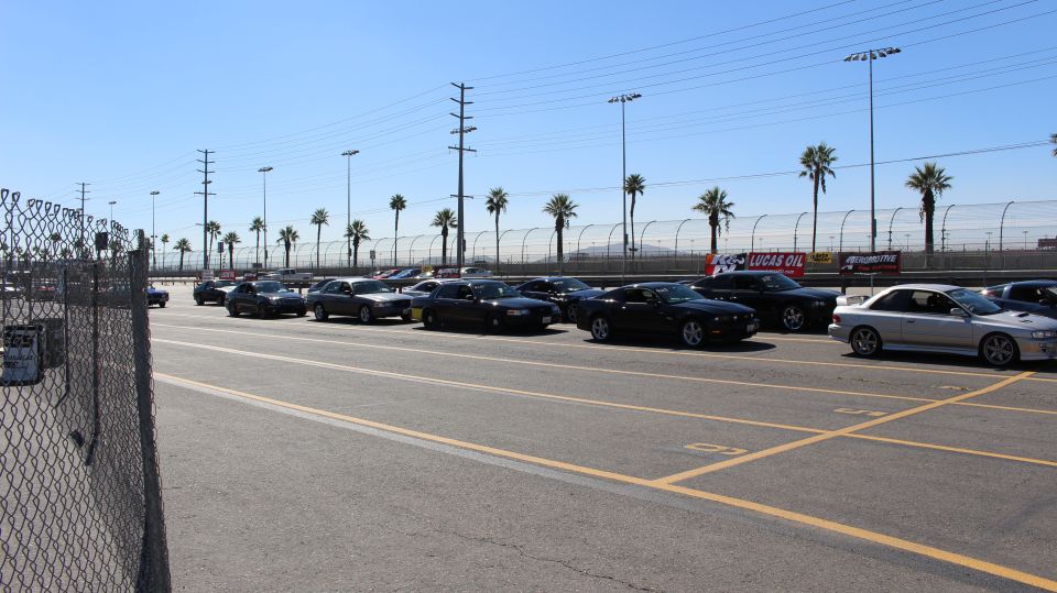 Line of dark coupes and sedans staged behind a chain-link fence at a track