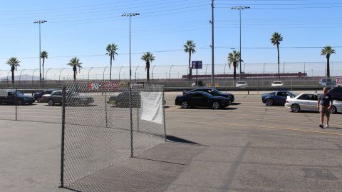 2003 Ford Crown Victoria - Coupes park in a paved track lot beyond a freestanding chain-link fence panel as a man stands at right under a clear sky.