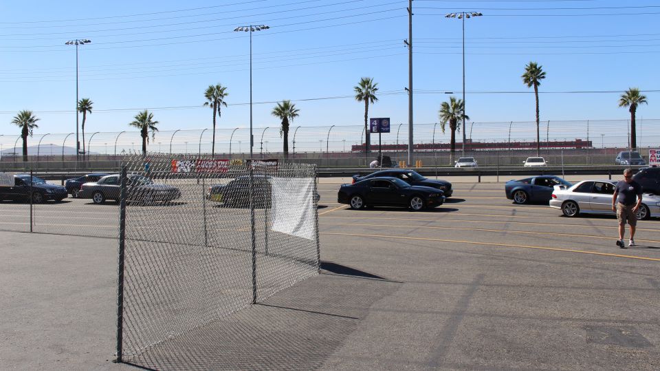 Coupes parked in a track lot beyond a chain-link fence panel and a standing man