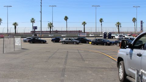 2003 Ford Crown Victoria - wide paved track lot holds scattered sedans and coupes, with a silver pickup parked in the foreground and palm trees beyond.
