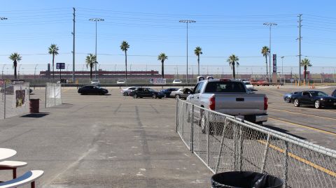2003 Ford Crown Victoria - silver pickup and several cars sit in a paved track lot behind metal fencing, with a trash barrel and palm trees nearby.