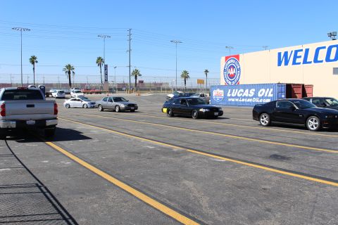 2003 Ford Crown Victoria - Sedans, a Ford pickup and coupes sit in a paved lot beside a building with "LUCAS OIL" and "WELCOME" signage under a clear sky.