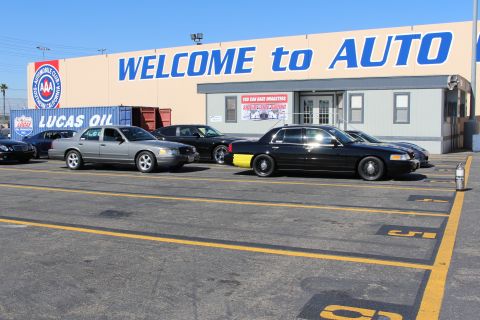 2003 Ford Crown Victoria - gray sedan and a black sedan parked in numbered lot spaces in front of a tan building reading WELCOME to AUTO with AAA and Lucas Oil signag…