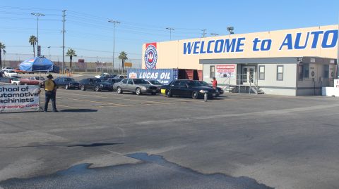 2003 Ford Crown Victoria - line of dark sedans parked along a tan building reading WELCOME to AUTO, with a blue canopy and a vendor banner at left on cracked pavement.