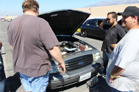 2003 Ford Crown Victoria - Several men stand around a gray Ford sedan with its hood raised, inspecting the engine bay, with mountains visible in the distance.