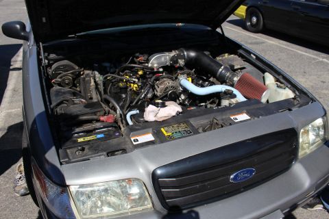 2003 Ford Crown Victoria - open engine bay of a gray Ford sedan showing a modified intake with a red cone air filter, black charge piping, and a clear coolant reservo…