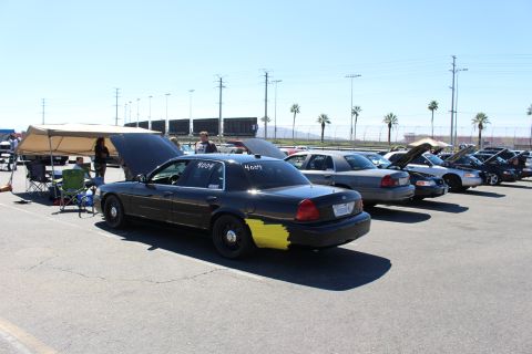 2003 Ford Crown Victoria - row of black and gray Ford Crown Victoria sedans parked at a track lot, with a canopy and chairs at left and grandstands behind.