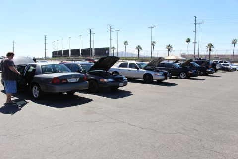 2003 Ford Crown Victoria - long row of Ford Crown Victoria sedans with hoods raised parked at a track lot, with a person tending an open trunk at left.