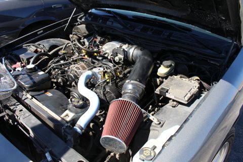 2003 Ford Crown Victoria - modified engine bay with a red cone air filter, polished aluminum intake piping and black charge tubing under a raised hood.
