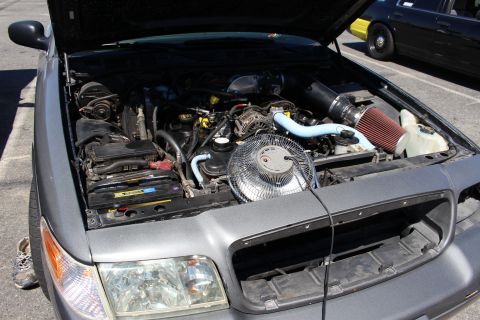 2003 Ford Crown Victoria - open engine bay of a gray sedan showing the mechanical cooling fan, blue coolant hoses and a red cone air filter.