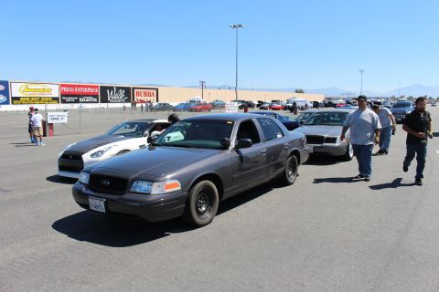 2003 Ford Crown Victoria - gray Ford Crown Victoria staged in a lineup of Crown Vics at a track, with people walking nearby and grandstand banners behind.