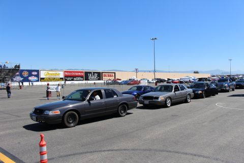 2003 Ford Crown Victoria - line of gray and dark Ford Crown Victoria sedans staged on track tarmac with a traffic cone in front and grandstand banners behind.