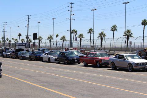 2003 Ford Crown Victoria - long line of vehicles, including a red classic coupe and silver coupe, waits in a sunny track lot beneath power lines and palm trees.