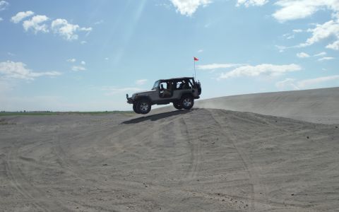 2005 Jeep Wrangler - A gray Jeep Wrangler takes flight over a sand dune, kicking up dust under a partly cloudy sky.