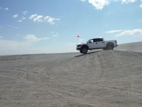2005 Jeep Wrangler - A white pickup truck drives on a sand dune under a blue sky with scattered clouds.
