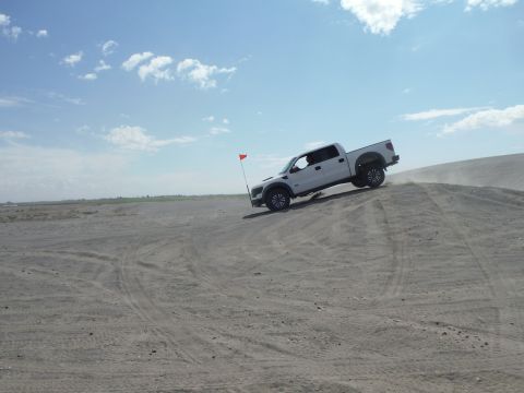 2005 Jeep Wrangler - White Ford F-150 Raptor navigates a sandy incline under a partly cloudy sky.