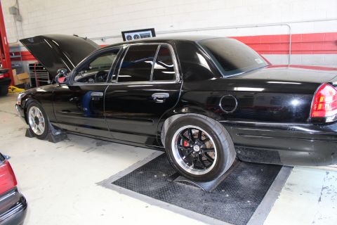 2003 Ford Crown Victoria - black full-size Ford sedan sits on shop rollers with its hood raised and staggered drag wheels against a red-and-white wall.