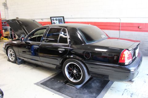2003 Ford Crown Victoria - Rear three-quarter view of a black full-size sedan on dyno rollers with wide rear tires inside a red-and-white shop.