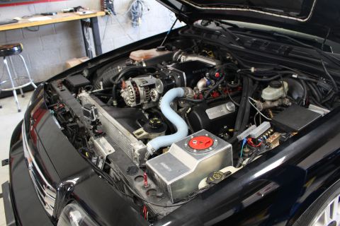 2003 Ford Crown Victoria - modified engine bay of a black car shows an aluminum intercooler tank with red cap and light-blue hose in a workshop.