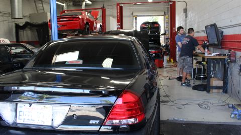 2003 Ford Crown Victoria - black Ford sedan badged 'SVT' sits in a service bay with technicians working and a red car raised on a lift behind it.