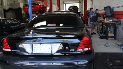 2003 Ford Crown Victoria - rear of a black Ford sedan with an 'SVT' badge and 'EVIL VIC' plate sits in a service bay with a red car on a lift behind.