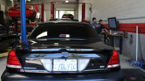 2003 Ford Crown Victoria - Black Ford sedan parked rear-first in a service garage, its California vanity plate reading EVIL VIC, with a red car on a lift behind it.