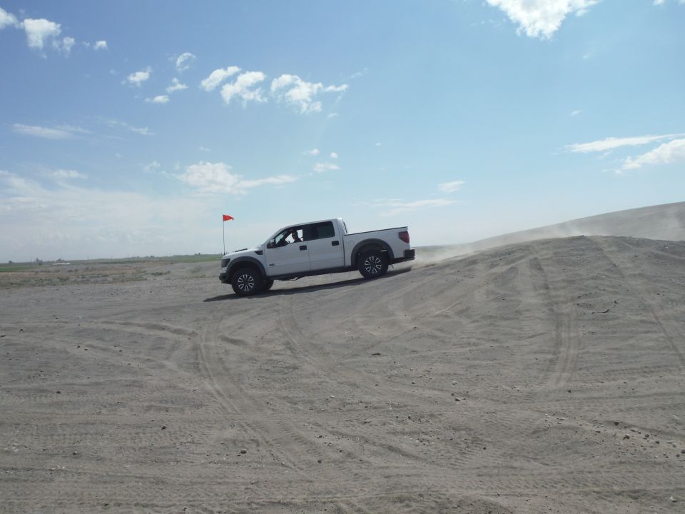 White pickup truck with black fender flares driving on a sand dune under a blue sky with clouds. A red flag is visible