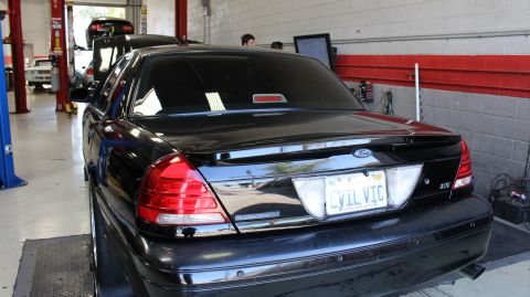 2003 Ford Crown Victoria - Black Ford sedan viewed from the rear three-quarter inside a repair shop, displaying a license plate that reads EVIL VIC.