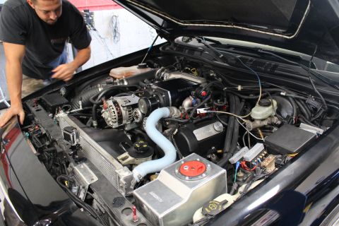 2003 Ford Crown Victoria - Open engine bay of a black car showing an alternator, blue intake tubing and turbo plumbing while a man works at the left.
