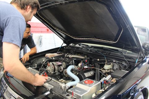 2003 Ford Crown Victoria - Two men lean over the open engine bay of a black car, inspecting the turbocharged motor with blue piping in a shop.