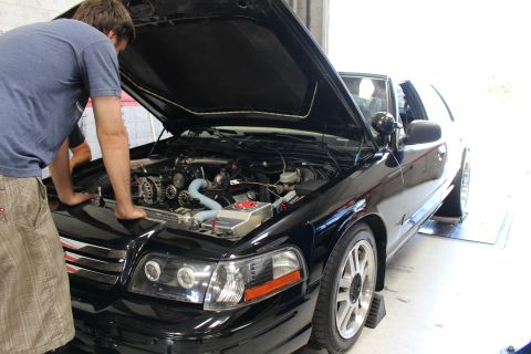 2003 Ford Crown Victoria - man reaches into the open engine bay of a black sedan parked on rollers in a garage, blue intake piping visible.