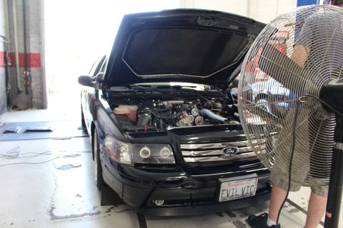 2003 Ford Crown Victoria - Black Ford sedan with hood up and turbo engine exposed sits beside a large industrial fan; front plate reads EVIL VIC.