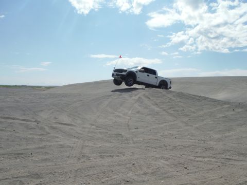 2005 Jeep Wrangler - A white pickup truck is captured mid-air as it jumps over a sand dune under a partly cloudy sky.