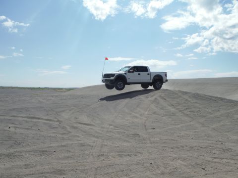 2005 Jeep Wrangler - A white pickup truck is captured mid-air as it jumps over a sand dune under a partly cloudy sky.