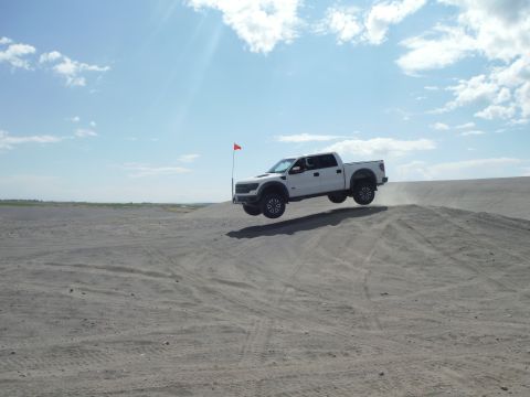 2005 Jeep Wrangler - A white pickup truck soars through the air above a sand dune under a partly cloudy sky.