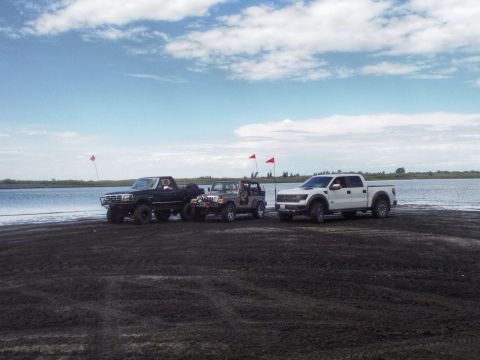2005 Jeep Wrangler - A black pickup, a gray Jeep, and a white Ford Raptor are parked together on a dark sandy area by the water under a cloudy sky.