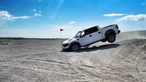 2005 Jeep Wrangler - A white pickup truck is captured mid-air as it drives over a sand dune under a bright blue sky.