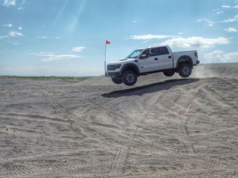 2005 Jeep Wrangler - A white pickup truck is captured mid-air as it jumps over a sand dune under a partly cloudy sky.