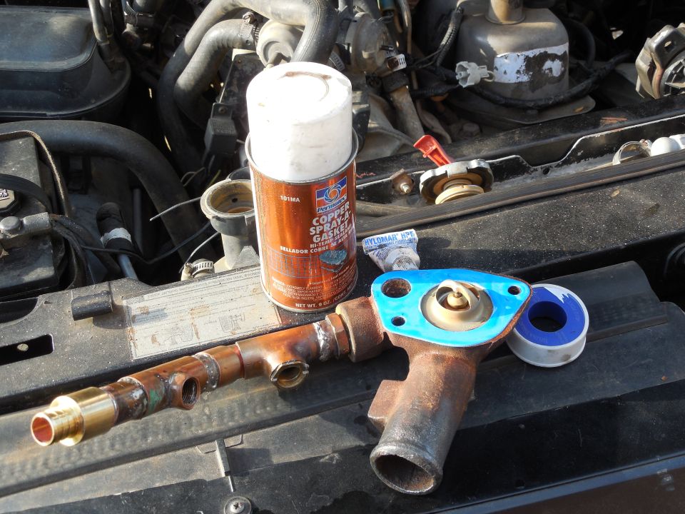 Rusty water neck with thermostat next to copper gasket spray, Hylomar sealant and PTFE tape on an engine.