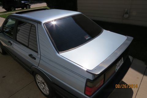 1990 Volkswagen Jetta - The rear quarter panel of a silver sedan is visible, parked in a driveway with a house in the background.