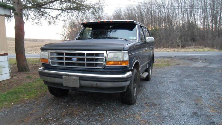 Dark blue full-size Ford Bronco parked on gravel, front three-quarter view with chrome grille and windshield visor