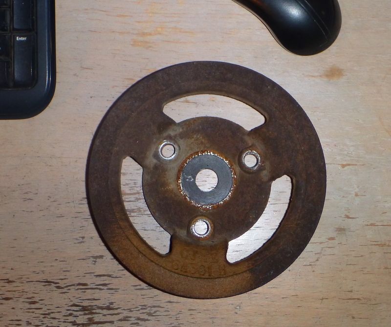 Rusty metal pulley with three large cutouts and three bolt holes, resting on a wooden surface next to a keyboard and mouse