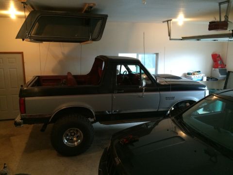 1989 Ford Bronco - A two-tone Ford Bronco with a removable hardtop stored overhead sits in a garage next to another vehicle.