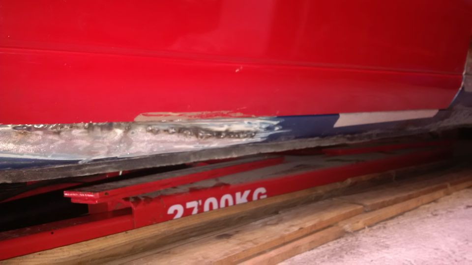 Red vehicle rocker panel shows rust and damage, possibly from an accident. It's on a lift with '2700KG' visible